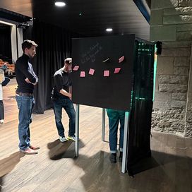 A few people standing around or in a large black box on stilts that has a few coloured post-its on its side